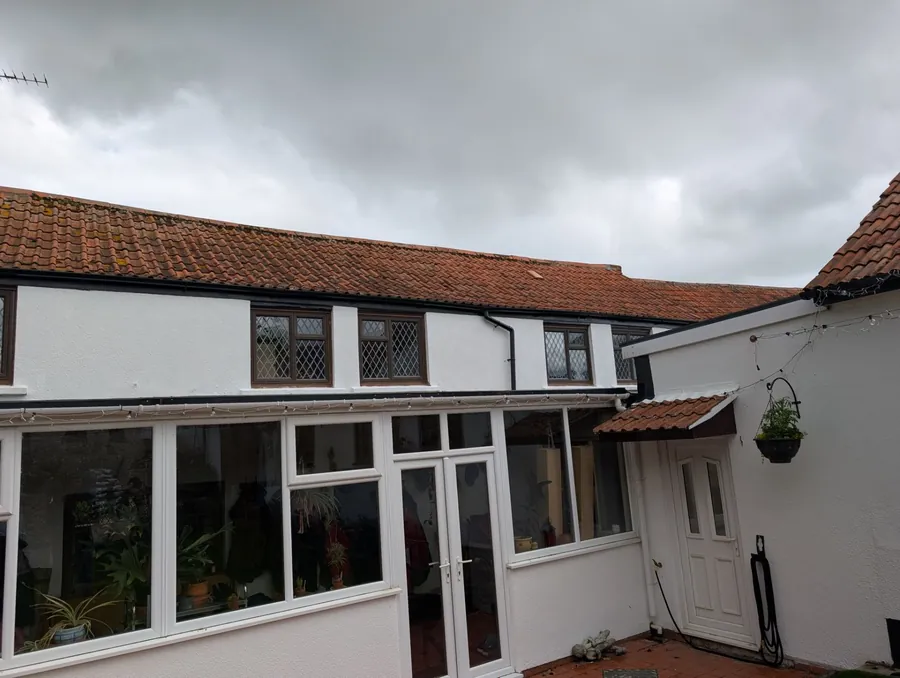 Rendered cottage with a pantile roof, leaded windows and a conservatory, photographed under a grey sky before roof repairs